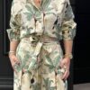 Une femme porte un ensemble chemise et short assortis, confectionné dans un tissu beige clair décoré de motifs tropicaux. Le design met en avant des palmiers verts, des toucans noirs, des léopards tachetés et des singes dorés, évoquant une ambiance exotique et élégante. La chemise, à manches longues retroussées, est rentrée dans le short, qui est ajusté à la taille grâce à une ceinture nouée dans le même tissu. Les accessoires dorés et un collier multicolore ajoutent une touche de raffinement à ce look décontracté et estival. L’arrière-plan noir met en valeur les tons naturels et vibrants de l’ensemble.