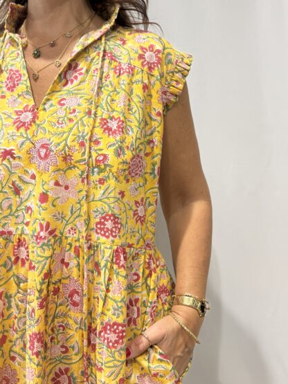 Robe Chandra Jaune - Femme portant une robe longue sans manches à imprimé floral, avec volants et liens à pompons, coupe ample et col tunisien.