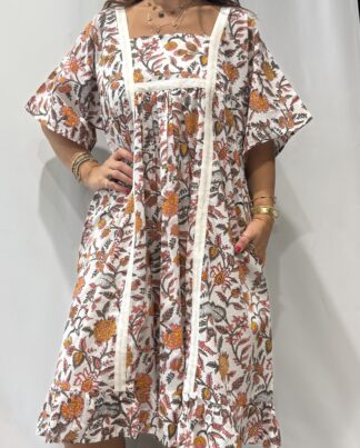 Robe Mallika Blanche Fleurie : Femme portant une robe blanche à imprimé floral rose orangé, avec empiècements contrastés blancs et manches courtes amples.
