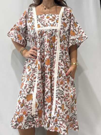 Robe Mallika Blanche Fleurie : Femme portant une robe blanche à imprimé floral rose orangé, avec empiècements contrastés blancs et manches courtes amples.