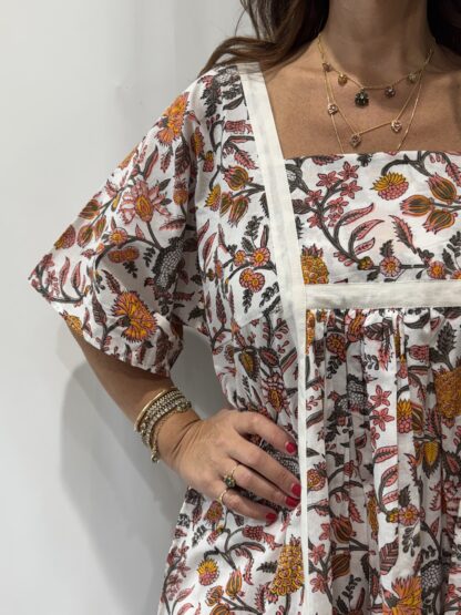 Robe Mallika Blanche Fleurie : Femme portant une robe blanche à imprimé floral rose orangé, avec empiècements contrastés blancs et manches courtes amples.