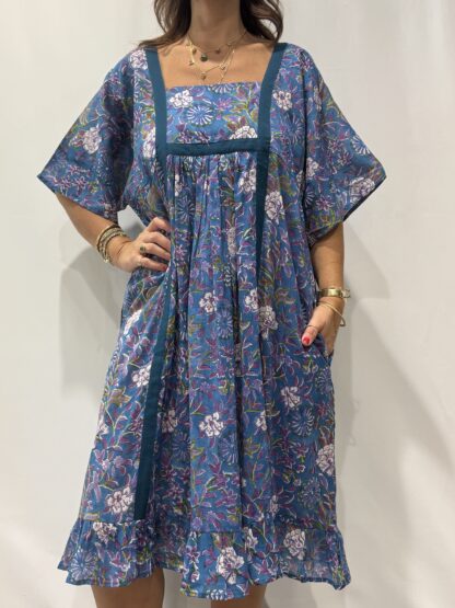 Robe Mallika bleu mauve : Femme portant une robe bleu à imprimé floral vert et violet avec empiècements contrastés bleu et manches courtes amples.