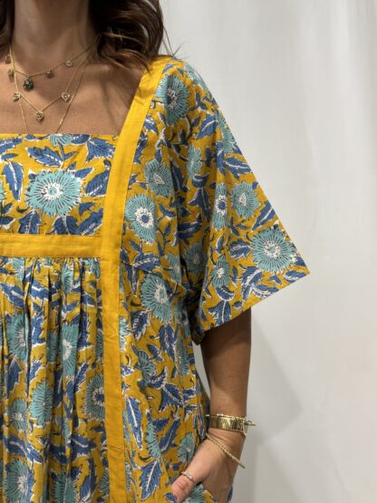 Robe Mallika Jaune Bleu : Femme portant une robe jaune à imprimé floral bleu avec empiècements contrastés jaune et manches courtes amples.