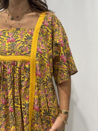 Robe Mallika Jaune Fleurie : Femme portant une robe jaune à imprimé floral vert et rose avec empiècements contrastés jaunes et manches courtes amples.