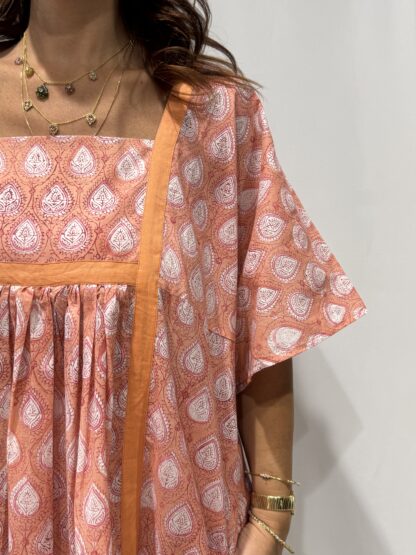 Robe Mallika Saumon : Femme portant une robe rose saumon à imprimé floral blanc avec empiècements contrastés rose saumon et manches courtes amples.