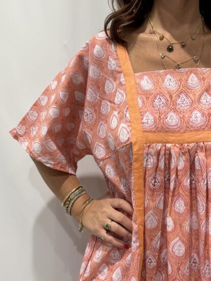 Robe Mallika Saumon : Femme portant une robe rose saumon à imprimé floral blanc avec empiècements contrastés rose saumon et manches courtes amples.