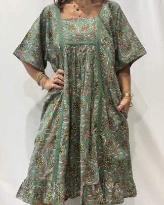 Robe Mallika Verte: Femme portant une robe verte à imprimé floral marron, jaune et bleu avec empiècements contrastés vert et manches courtes amples.
