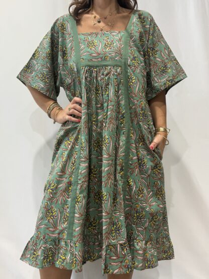 Robe Mallika Verte: Femme portant une robe verte à imprimé floral marron, jaune et bleu avec empiècements contrastés vert et manches courtes amples.