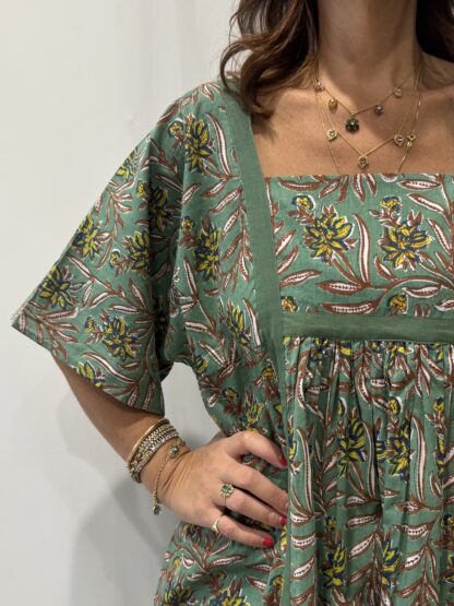 Robe Mallika Verte: Femme portant une robe verte à imprimé floral marron, jaune et bleu avec empiècements contrastés vert et manches courtes amples.