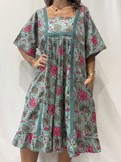 Robe Mallika Verte Rose : Femme portant une robe verte à imprimé floral rose et vert, avec empiècements contrastés jaunes et manches courtes amples.