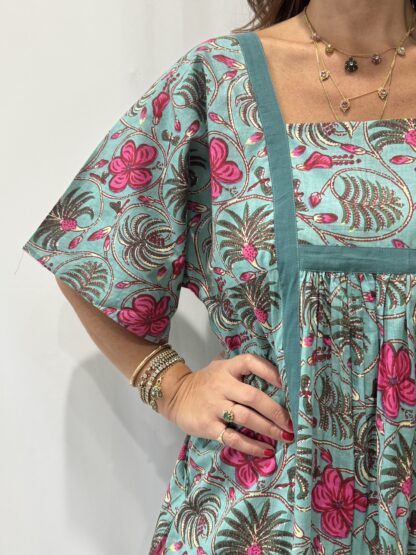 Robe Mallika Verte Rose : Femme portant une robe verte à imprimé floral rose et vert, avec empiècements contrastés jaunes et manches courtes amples.