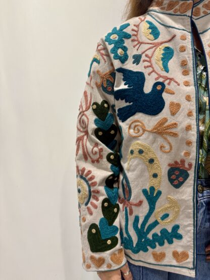 Veste Namita de Micasasté pour femme, brodée sur fond clair avec motifs colorés bleu, vert, orangé, jaune pâle et vieux rose, comprenant des fleurs, des cœurs et des oiseaux.