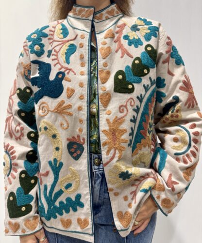 Veste Namita de Micasasté pour femme, brodée sur fond clair avec motifs colorés bleu, vert, orangé, jaune pâle et vieux rose, comprenant des fleurs, des cœurs et des oiseaux.