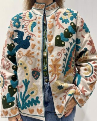 Veste Namita de Micasasté pour femme, brodée sur fond clair avec motifs colorés bleu, vert, orangé, jaune pâle et vieux rose, comprenant des fleurs, des cœurs et des oiseaux.