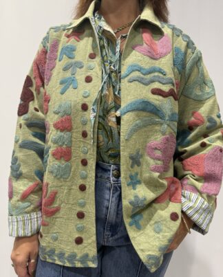 Veste Veda de Micasasté, portée de face, aux broderies florales rose, mauve, marron, bleu et bleu-gris sur fond vert. Une veste courte aux manches longues et col mao. Une veste Suzani embrodery.
