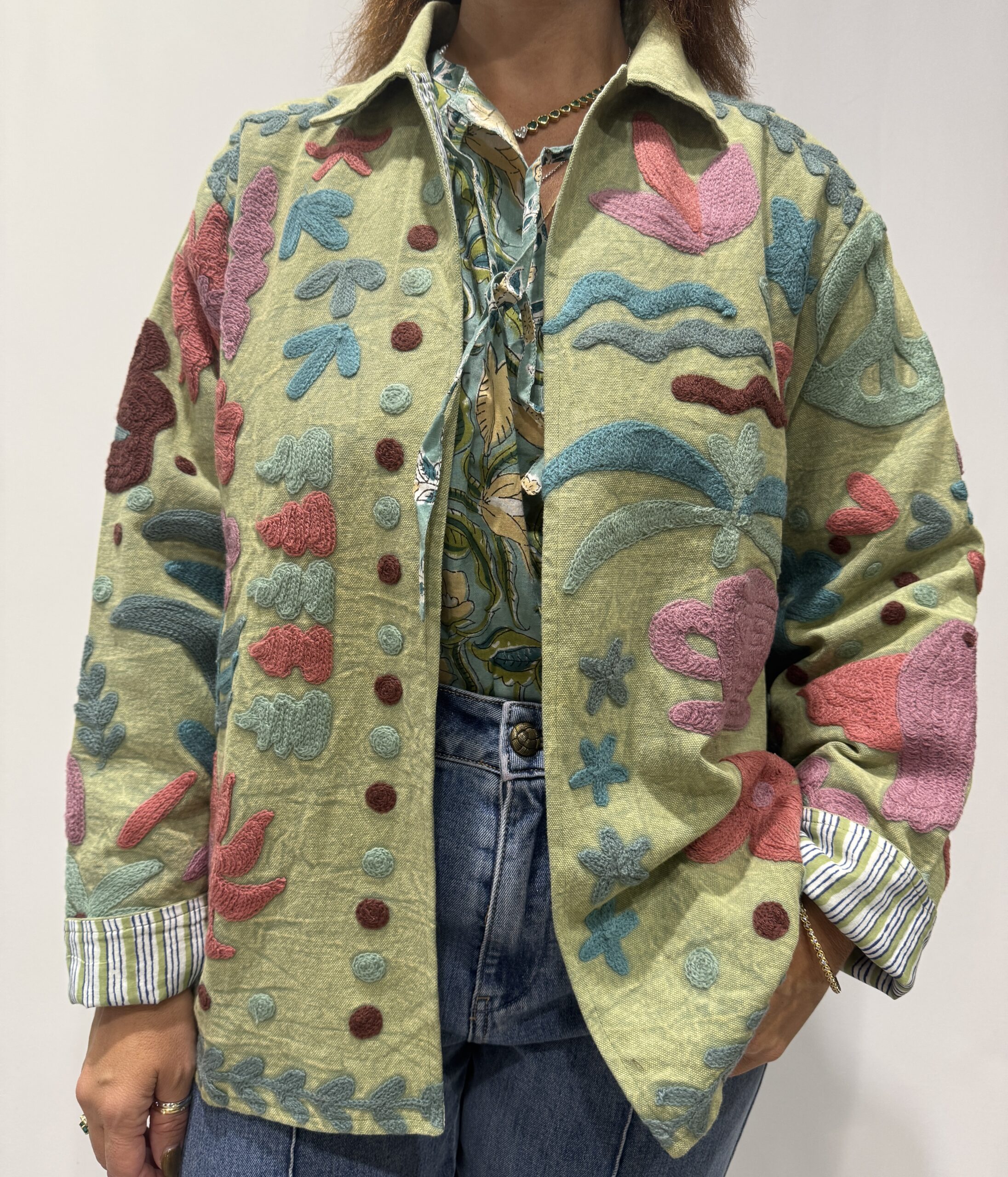 Veste Veda de Micasasté, portée de face, aux broderies florales rose, mauve, marron, bleu et bleu-gris sur fond vert. Une veste courte aux manches longues et col mao. Une veste Suzani embrodery.