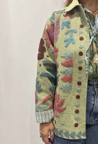 Veste Veda de Micasasté portée de profil droit, aux broderies colorées sur fond vert. Une veste courte suzani embrodery aux manches longues et col mao.