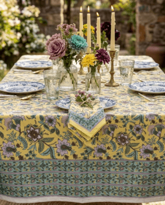 Nappe Peela à fond jaune lumineux et motifs floraux violets, avec une frise décorative verte et mauve sur les bordures. La table est dressée en extérieur avec de la vaisselle raffinée, des bougies et des bouquets de fleurs colorées.