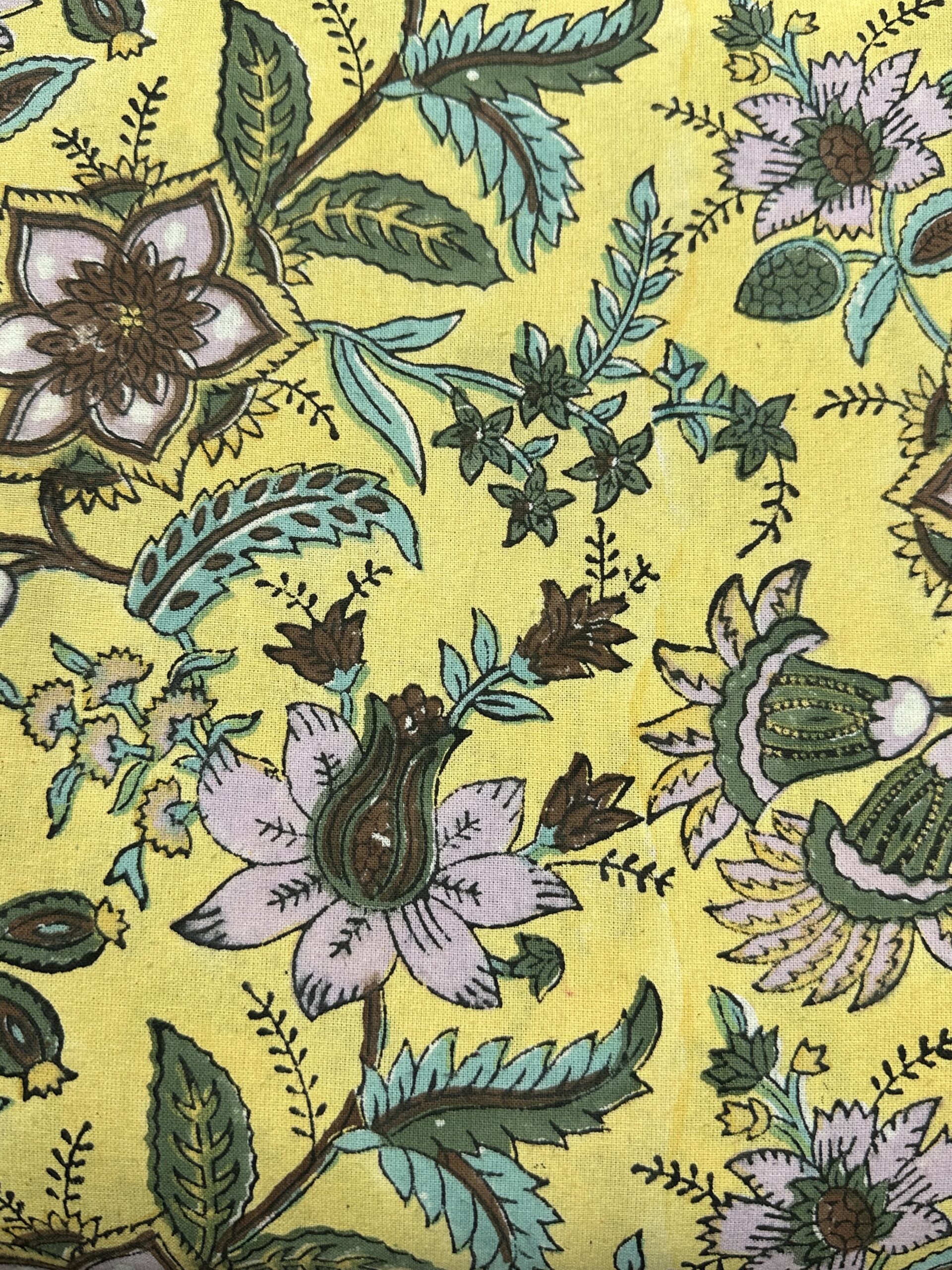 Nappe Peela à fond jaune lumineux et motifs floraux violets, avec une frise décorative verte et mauve sur les bordures. La table est dressée en extérieur avec de la vaisselle raffinée, des bougies et des bouquets de fleurs colorées.